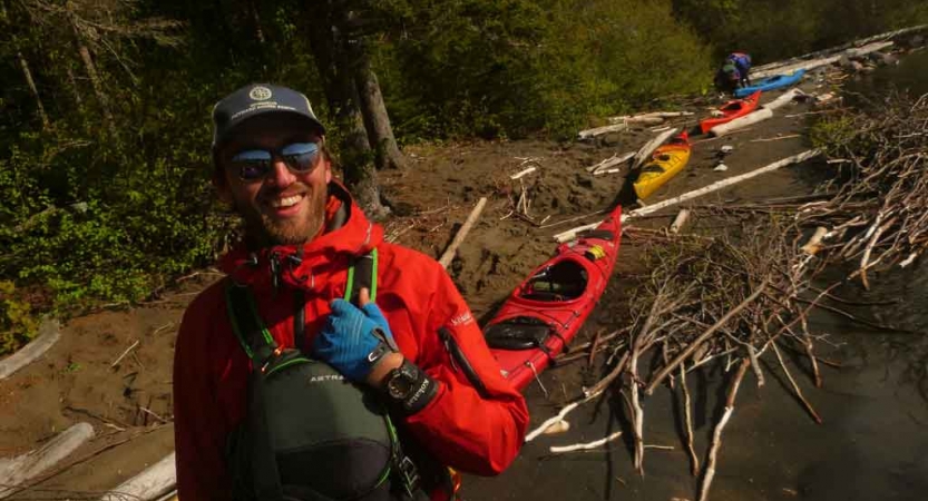 a student smiles at the camera with kayaks resting on the shore in the background